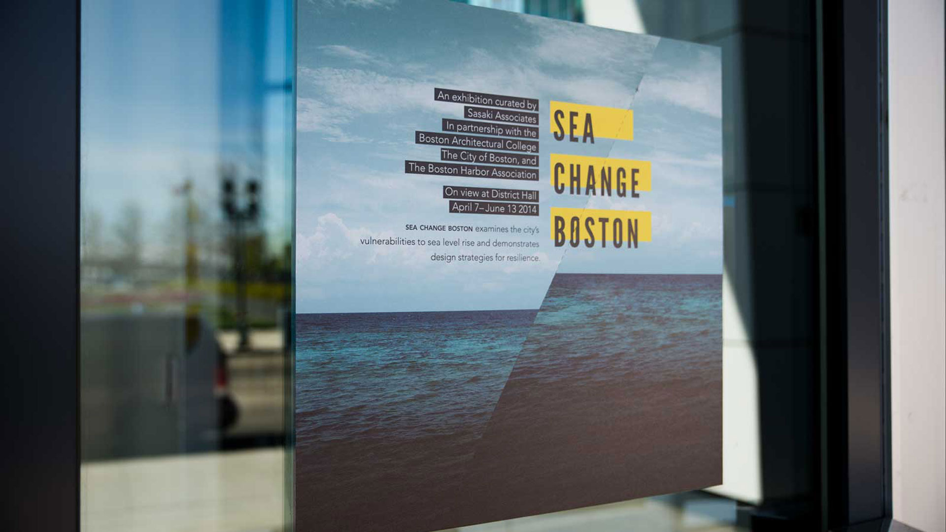 A photogarph of a sign on a window. The sign has a photo of the ocean on it with a dividing line and the ocean shifted up on the right side. There is also typogarphy and yellow horizontal bars on the sign. Design made in Adobe Indesign, Adobe Illustrator, and Adobe Photoshop.